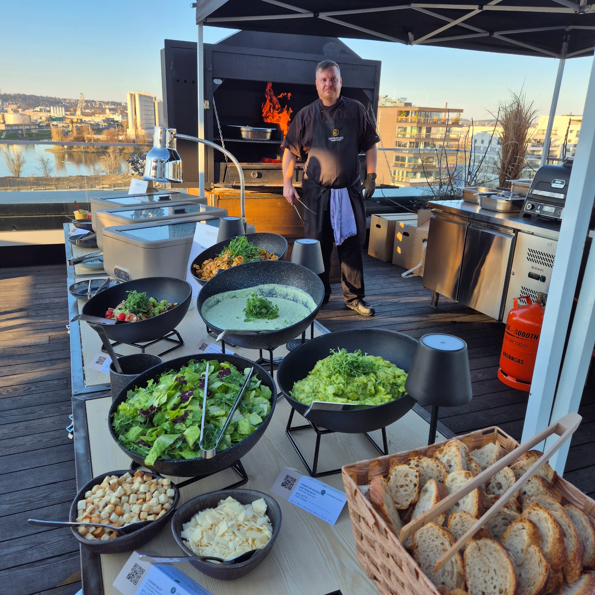 Grillstation mit Salaten, Beilagen und Brot auf einer Dachterrasse, dahinter ein Koch am offenen Grill